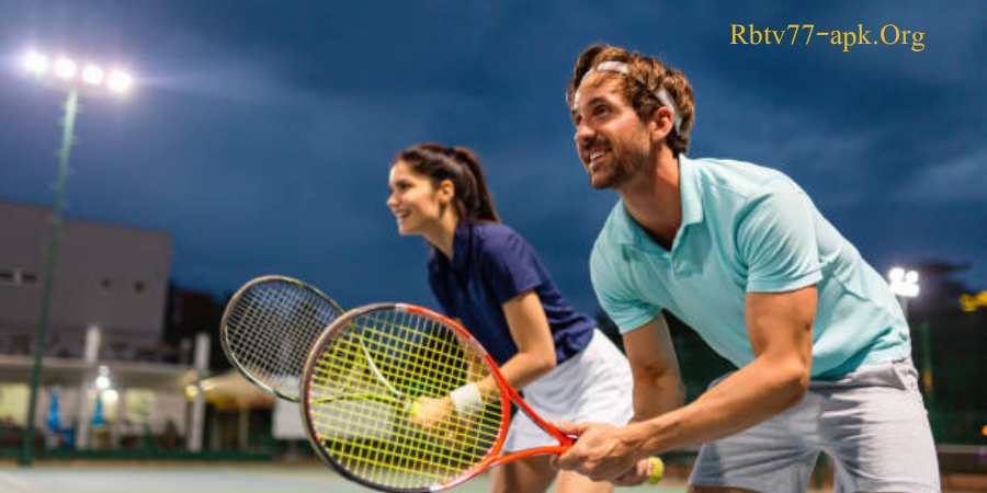 Smiling man and woman playing doubles tennis at night under stadium lights, both holding rackets and ready for action — Rbtv77-apk.org.